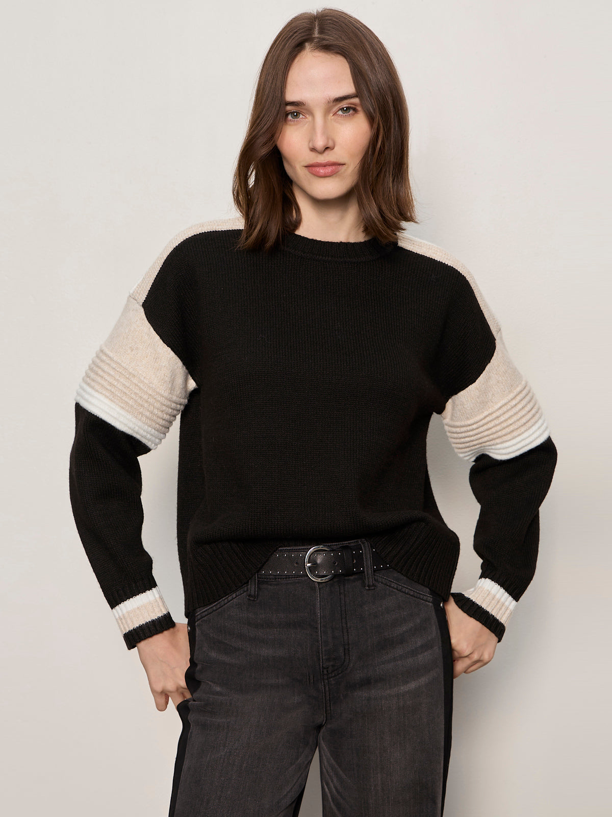 A woman with straight brown hair models the Sanctuary Clothing sporty stripe crewneck sweater in black multi, featuring beige and white sleeve stripes. She pairs it with dark high-waisted jeans and stands against a light background, looking at the camera.