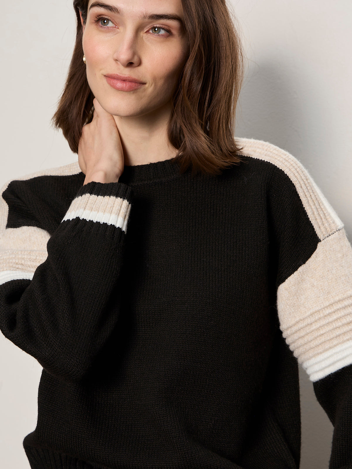 A woman with shoulder-length brown hair wears the Sanctuary Clothing sporty stripe crewneck sweater in black multi, touching her neck with one hand and smiling softly as she gazes slightly to the side.