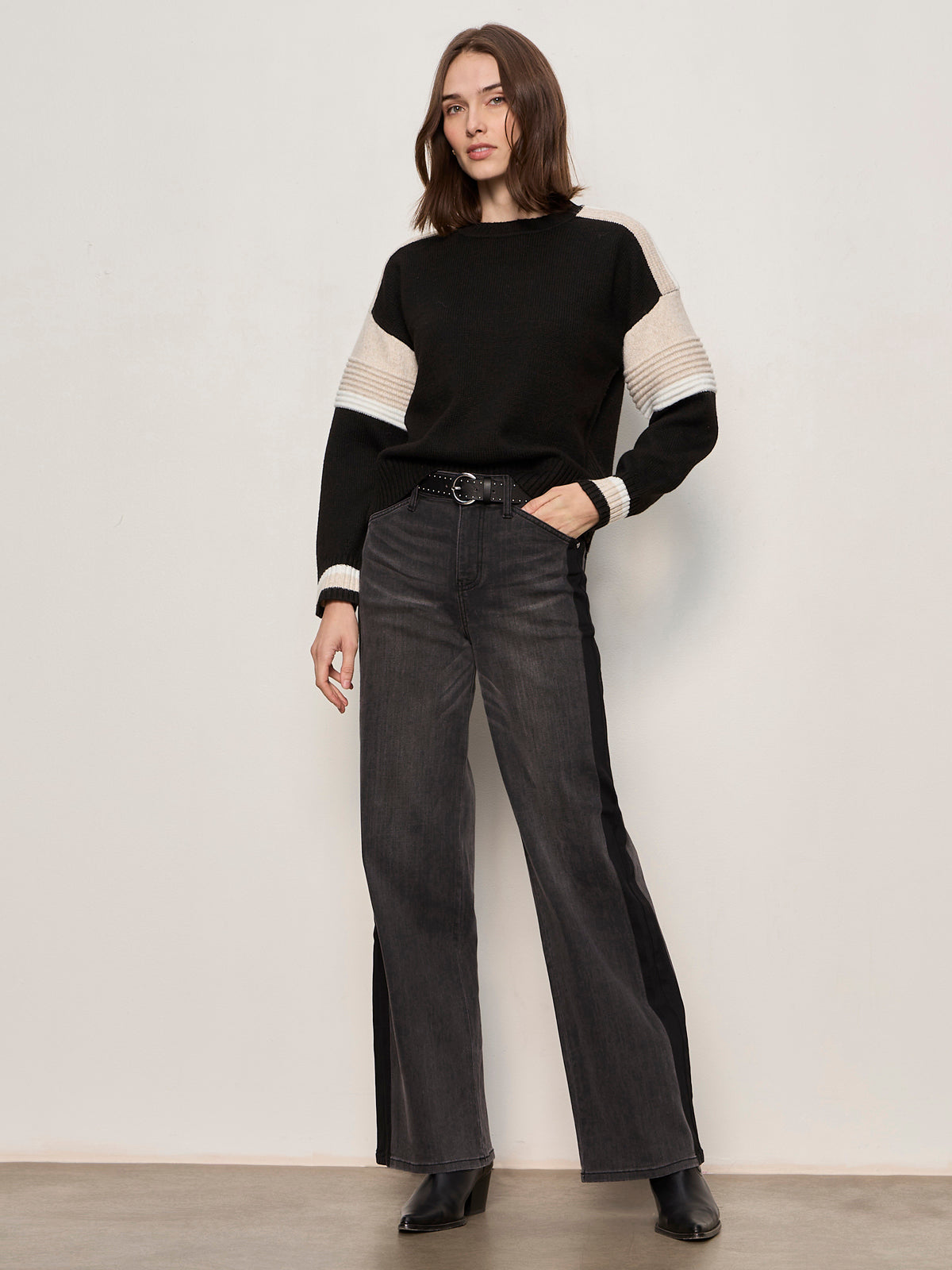 A woman with shoulder-length brown hair poses against a light wall, wearing the Sanctuary Clothing sporty stripe crewneck sweater in black multi, wide-leg dark gray jeans, and black shoes.