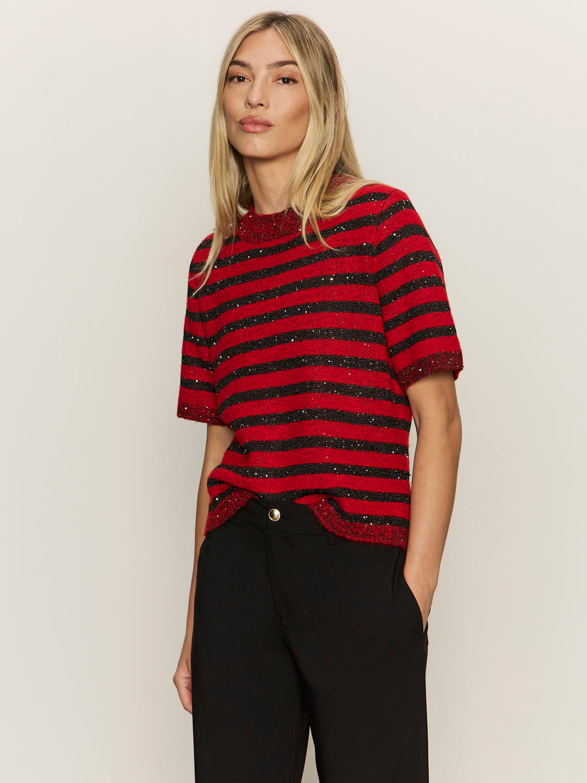 A woman with straight blonde hair wears the Sanctuary Clothing short sleeve shine sweater tee in cherry lacquer shine and black pants, standing against a plain light background with her hands in her pockets.