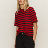 A woman with straight blonde hair wears the Sanctuary Clothing short sleeve shine sweater tee in cherry lacquer shine and black pants, standing against a plain light background with her hands in her pockets.