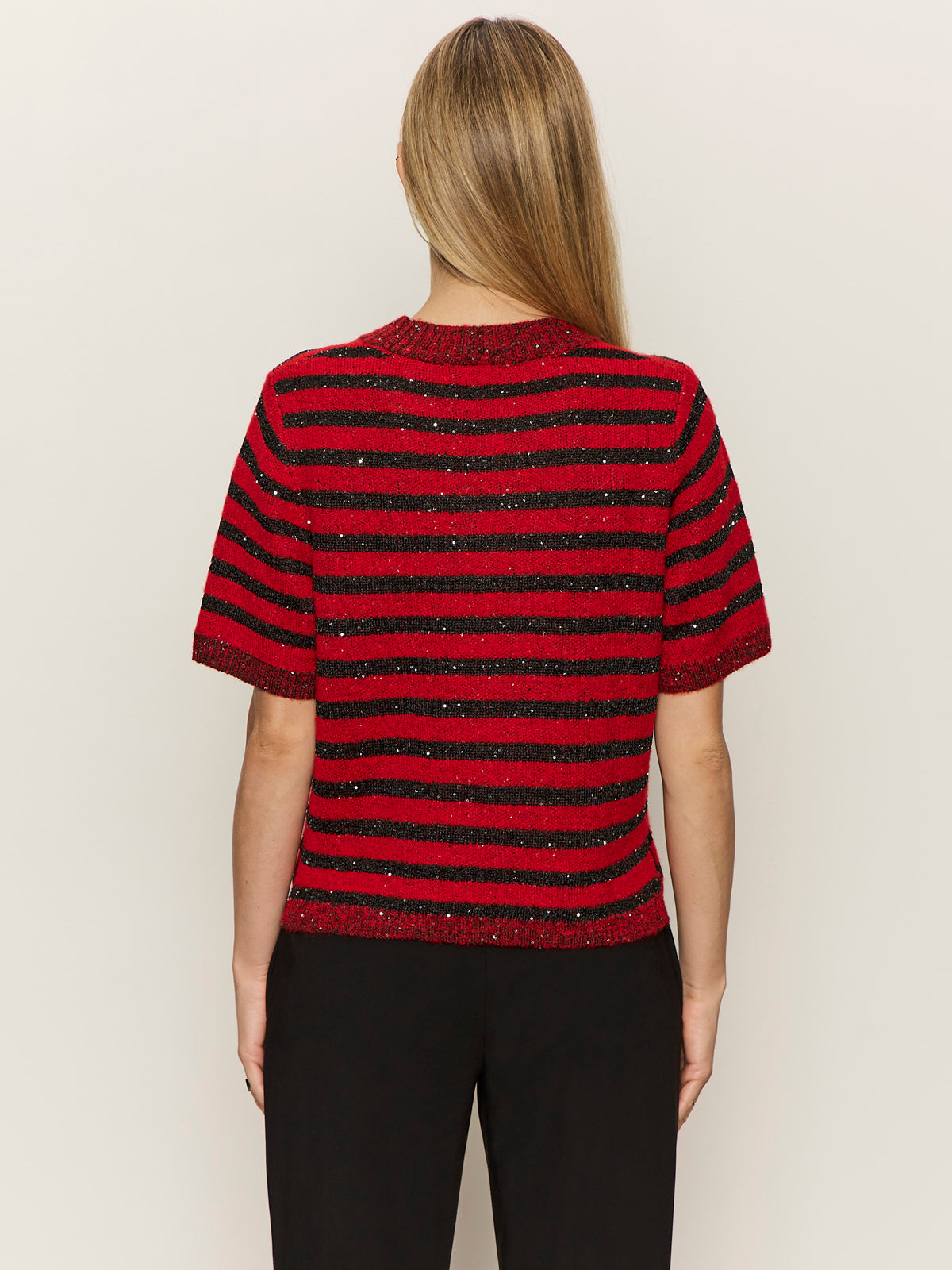 A person with long, straight blonde hair is seen from the back wearing the Sanctuary Clothing short sleeve shine sweater tee in cherry lacquer shine, paired with black pants, against a plain light background.