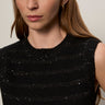 A woman with long brown hair wears the Sanctuary Clothing shine sweater shell in black with black shine, a sleeveless textured top featuring subtle sparkly stripes. She accessorizes with gold hoop earrings and faces the camera neutrally.