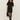 A woman with curly hair wears the Sanctuary Clothing shine t shirt sweater dress in black with black shine, styled with sheer tights, heeled boots, and a black handbag, standing against a light background.