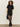 A woman with curly hair wears the Sanctuary Clothing shine t shirt sweater dress in black with black shine, styled with sheer tights, heeled boots, and a black handbag, standing against a light background.