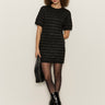 A woman with curly hair wears the Sanctuary Clothing shine t shirt sweater dress in black with black shine, styled with sheer tights, heeled boots, and a black handbag, standing against a light background.