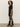 A woman with curly hair stands sideways, wearing the Sanctuary Clothing shine t shirt sweater dress in black with black shine, paired with sheer black tights and shiny black mid-calf boots against a plain light wall.