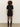 A person with curly hair faces away, wearing the Sanctuary Clothing shine t shirt sweater dress in black with black shine, paired with semi-sheer tights and knee-high boots, against a plain light wall and floor.