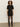 A woman with curly hair stands against a plain background, wearing the Shine T Shirt Sweater Dress in black with black shine by Sanctuary Clothing, paired with black sheer tights and black knee-high boots.
