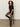 A woman with curly hair sits on a tall stool against a light wall, wearing Sanctuary Clothing's shine t shirt sweater dress black with black shine. She looks calmly at the camera, hands resting on her knees.