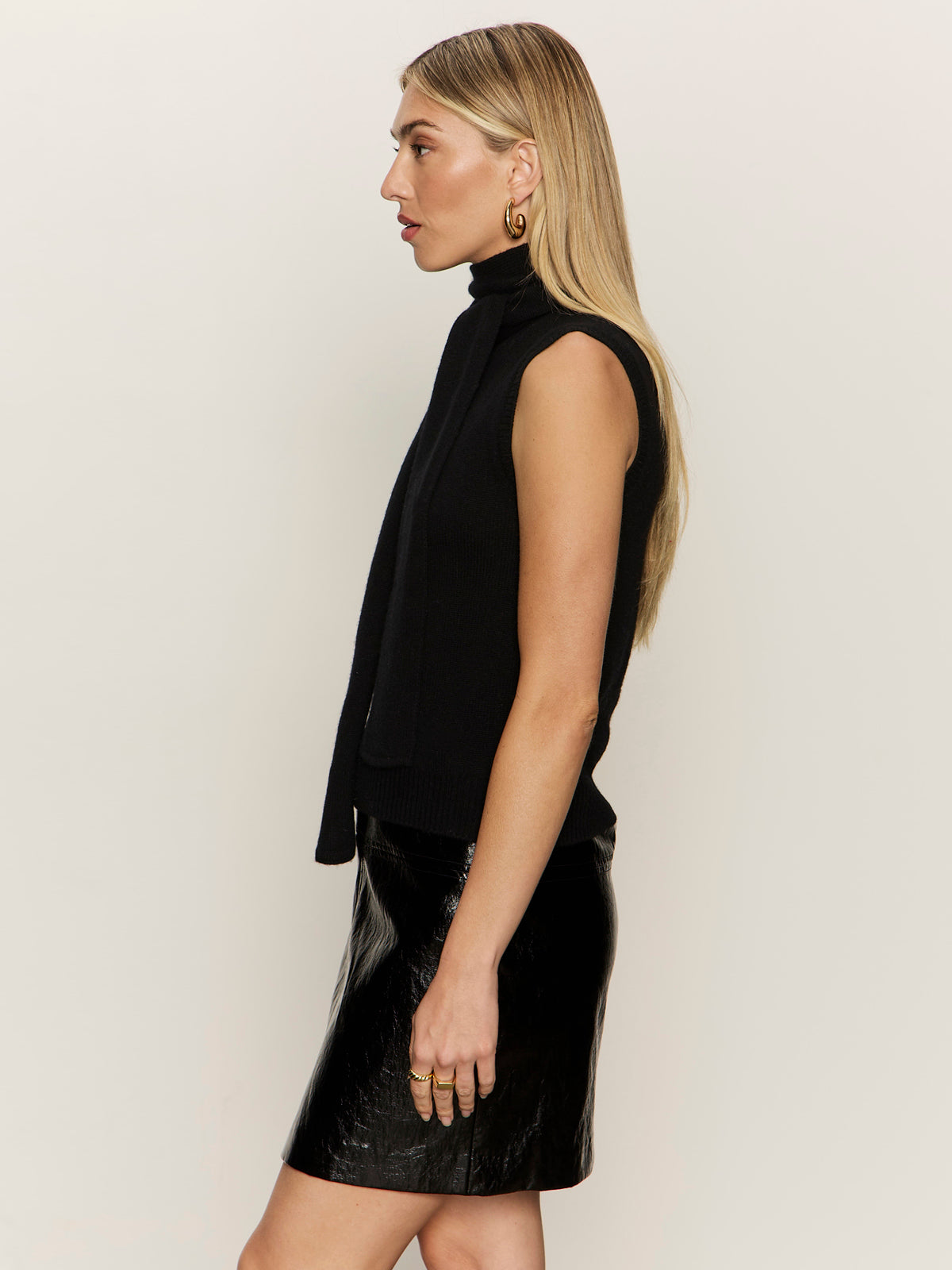 A woman with long blonde hair stands in profile against a plain light background, wearing the Sanctuary Clothing scarf sweater shell in black and gold hoop earrings, with rings on her fingers.
