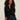 A woman with curly hair models the Sanctuary Clothing must have gem cardi black, a button-up sweater with small floral accents, paired with high-waisted dark blue jeans against a light-colored wall.