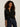 A woman with curly hair models the Sanctuary Clothing must have gem cardi black, a button-up sweater with small floral accents, paired with high-waisted dark blue jeans against a light-colored wall.
