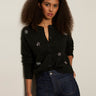 A woman with curly hair models the Sanctuary Clothing must have gem cardi black, a button-up sweater with small floral accents, paired with high-waisted dark blue jeans against a light-colored wall.