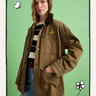 A woman stands against a green background wearing Sanctuary Clothing's "save our planet jacket timber," featuring cartoon patches, paired with a black and white striped shirt and blue jeans. Hand-drawn doodles decorate the image's border.