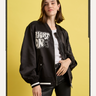 A woman with shoulder-length brown hair stands against a yellow background, wearing the Sanctuary Clothing right on jacket black over a white t-shirt and black pants, her expression neutral.