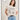 A woman in jeans and a pink "arent gonna keep quiet tee milk" by Sanctuary Clothing, featuring a cartoon print, stands smiling with one hand in her pocket against a white background with cloud doodles.