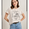 A woman stands with her hands on her hips, wearing light blue jeans and the Sanctuary Clothing "the doctor is in tee collagen," a white T-shirt with a Peanuts cartoon of Lucy at her "Psychiatric Help 5¢. The Doctor is IN" booth.