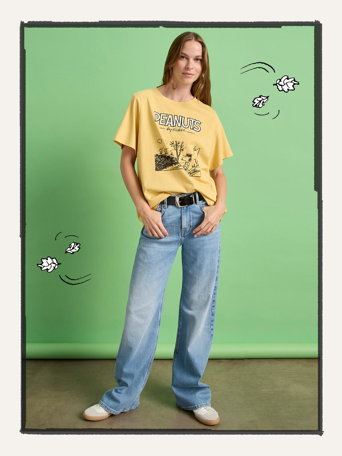 A woman stands against a green backdrop wearing the Sanctuary Clothing marching tee in washed gold, light blue wide-leg jeans, and white sneakers. Illustrated white leaves and wind lines surround her.