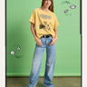 A woman stands against a green backdrop wearing the Sanctuary Clothing marching tee in washed gold, light blue wide-leg jeans, and white sneakers. Illustrated white leaves and wind lines surround her.