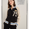 A woman with shoulder-length brown hair smiles with her hands in her pockets, wearing the Sanctuary Clothing 75 bomber sweater in black chalk. "STRIKE ONE!" appears beside her.