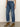 A person wearing Sanctuary Clothing's soleil 90s straight jeans in indigo medium, with rolled-up cuffs and brown heeled ankle boots, stands on a light brown floor against a neutral background.