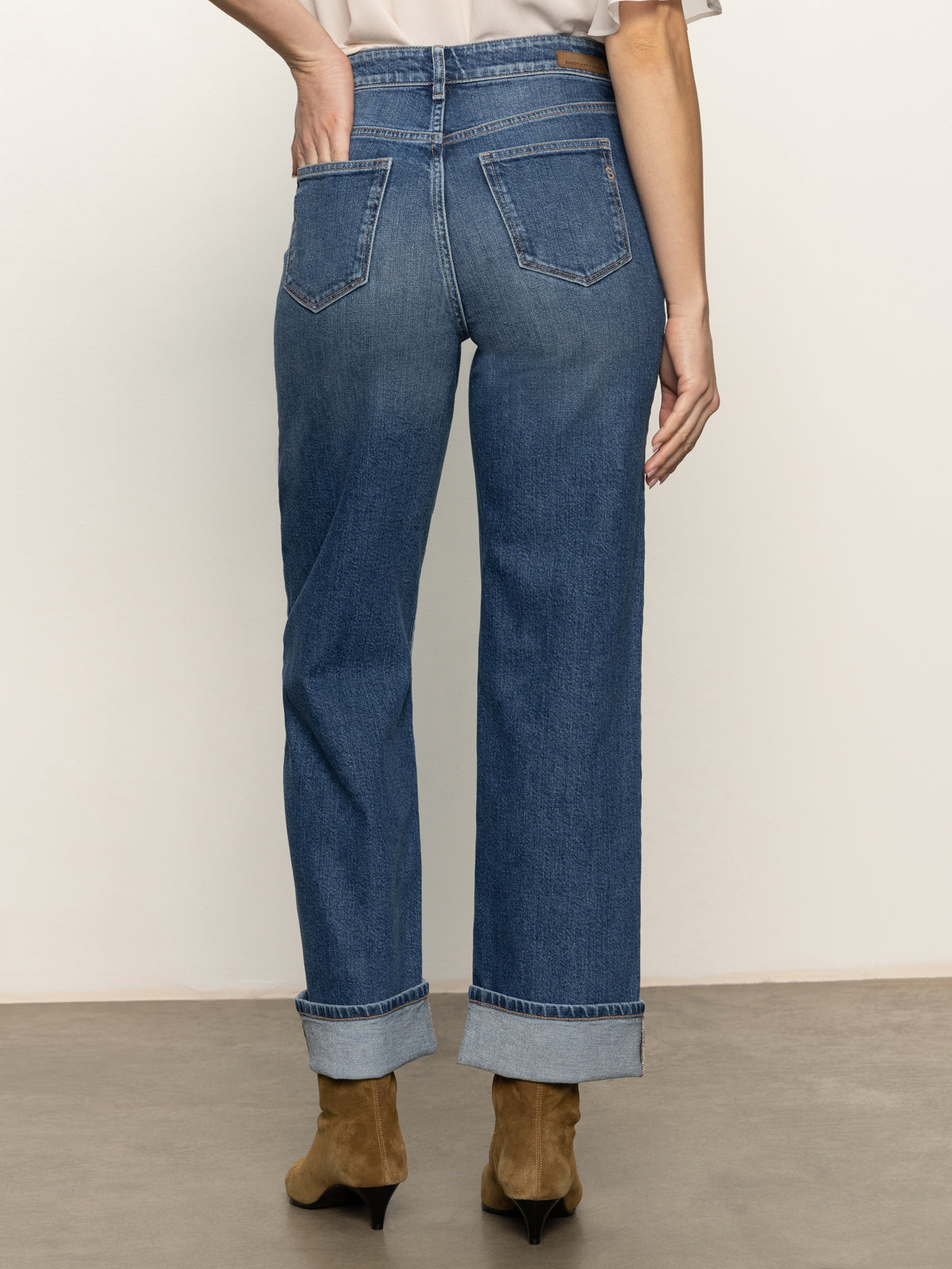 A person stands on a beige floor facing away, wearing medium indigo soleil 90s straight jeans by Sanctuary Clothing, with cuffed hems, brown ankle boots, and a light shirt. One hand rests on their hip.