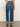 A person stands on a beige floor facing away, wearing medium indigo soleil 90s straight jeans by Sanctuary Clothing, with cuffed hems, brown ankle boots, and a light shirt. One hand rests on their hip.