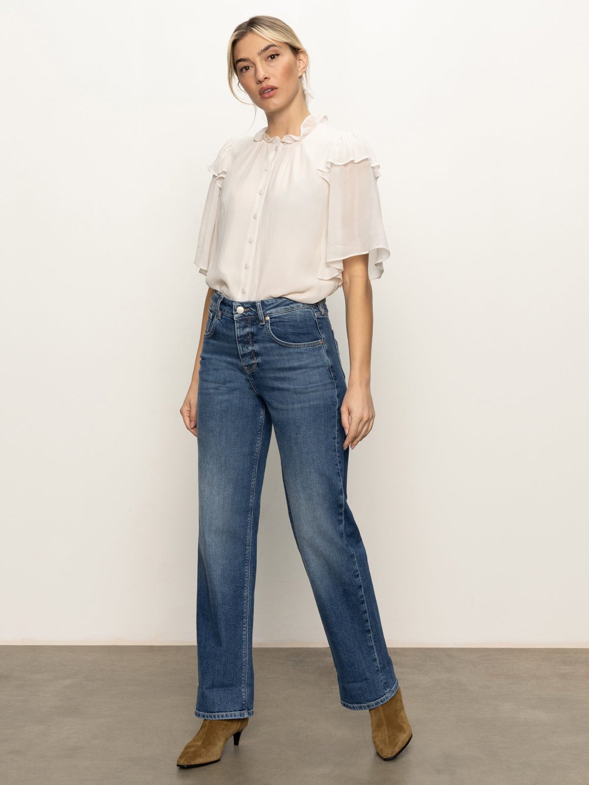 A woman with blonde hair tied back stands confidently against a plain background wearing Sanctuary Clothing's soleil 90s straight jeans in indigo medium, paired with a white ruffled blouse and tan heeled boots.