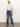 A woman with blonde hair tied back stands confidently against a plain background wearing Sanctuary Clothing's soleil 90s straight jeans in indigo medium, paired with a white ruffled blouse and tan heeled boots.