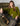 A woman with long brown hair sits confidently on a green velvet armchair, wearing the Sanctuary Clothing must have cardi martini olive, paired with dark pants and brown boots, her leg casually draped over the armrest.