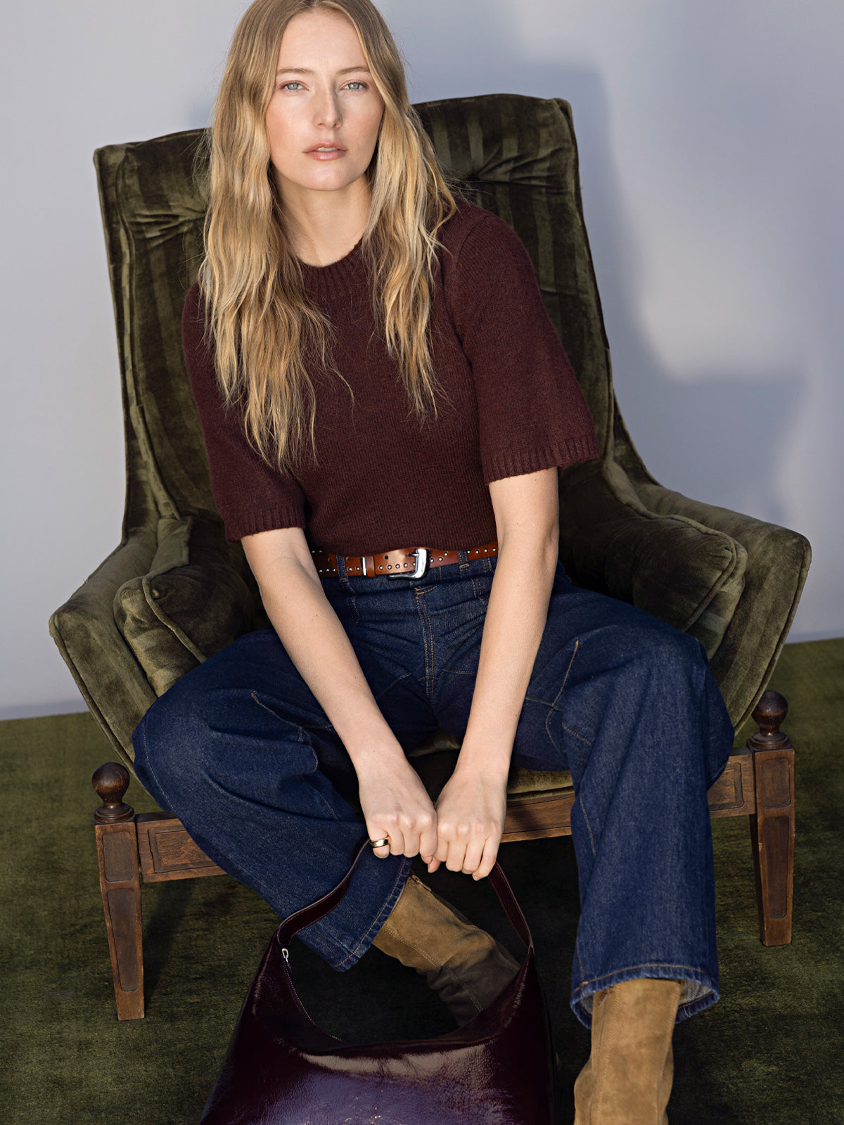 A woman with long blonde hair sits on a dark green armchair, wearing Sanctuary Clothing's perfect sweater tee in chicory, blue wide-leg jeans, a brown belt, tan suede boots, and holding a dark brown handbag.