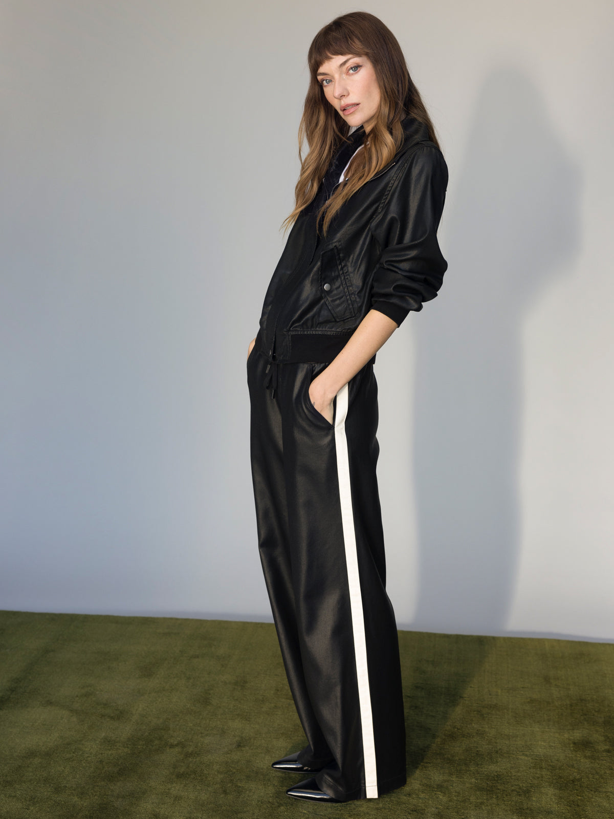 A woman stands on a green carpet, hands in pockets, wearing Sanctuary Clothing’s coated twill track pant in black with white side stripes and a black leather jacket, looking confidently at the camera against a plain light background.