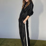 A woman stands on a green carpet, hands in pockets, wearing Sanctuary Clothing’s coated twill track pant in black with white side stripes and a black leather jacket, looking confidently at the camera against a plain light background.