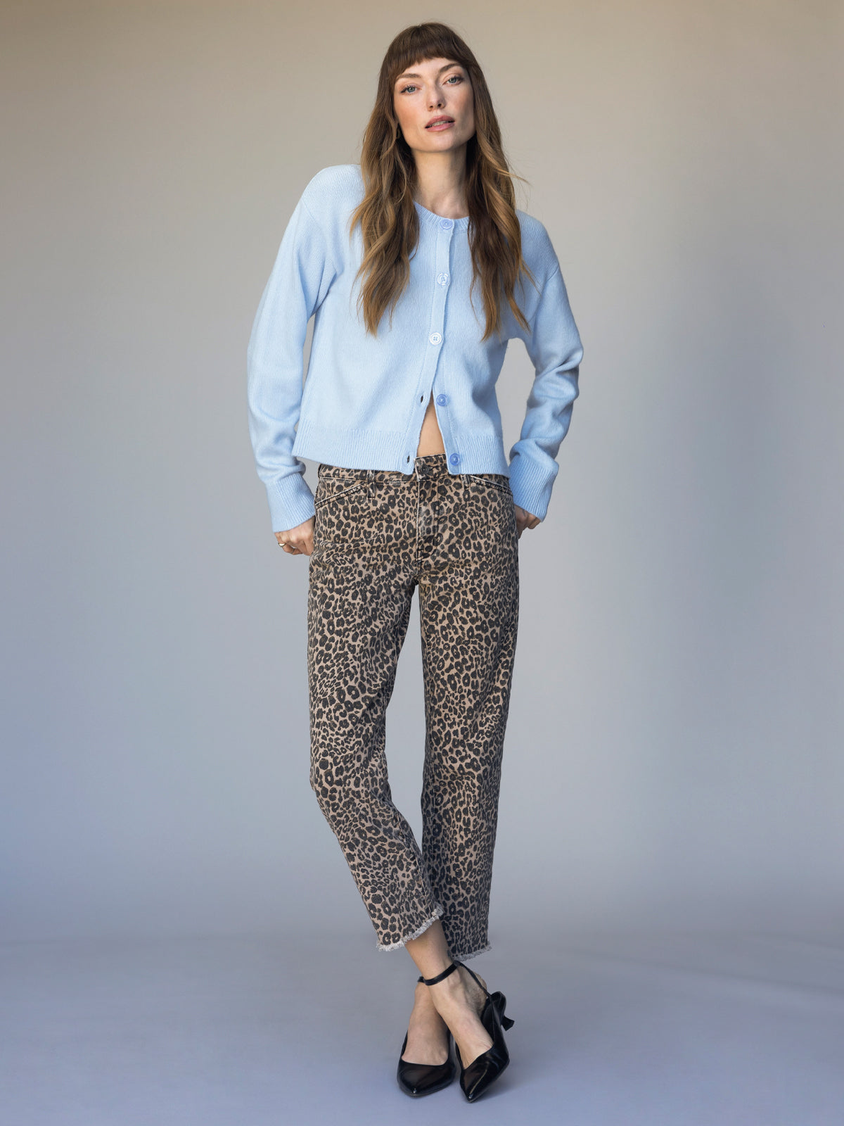 A woman with long brown hair and bangs stands against a plain background, wearing the Sanctuary Clothing must have cardi blue powder, leopard print pants, and black heels, looking at the camera with a relaxed posture.
