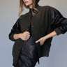 A woman poses against a plain background in the Sanctuary Clothing club varsity jacket in midnight olive, styled with a black leather mini skirt. She has long, wavy hair and looks to the side with one hand on her waist.
