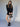 A woman stands against a plain background wearing Sanctuary Clothing's quilted velvet mini skirt in black, paired with a matching velvet jacket, sheer-detailed black ankle socks, black heeled shoes, and a small black polka dot handbag.