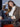 A woman with long brown hair sits on a red chair, confidently posing in the Sanctuary Clothing hometown jacket lynx over a white blouse, styled with a black belt and dark jeans.