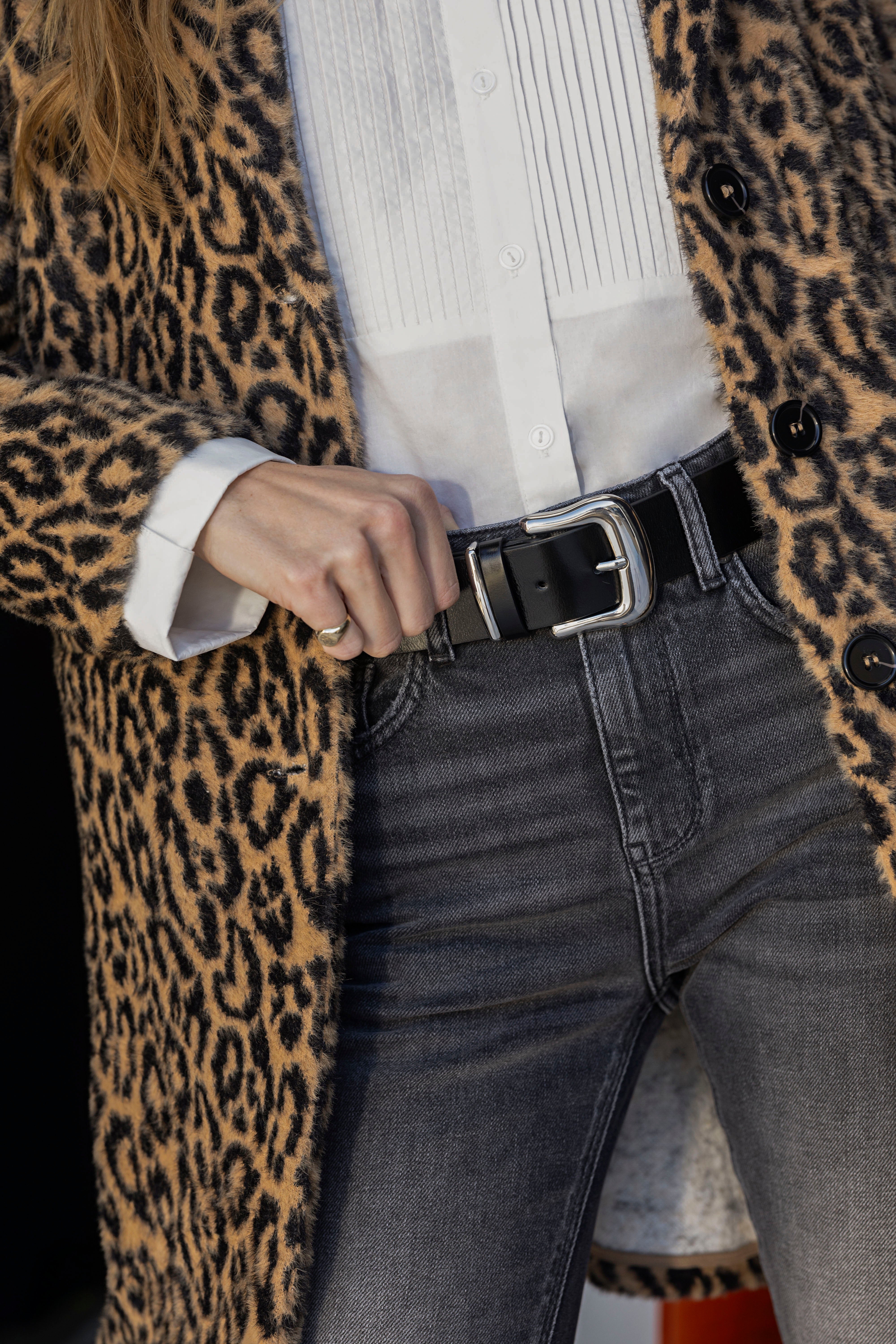 A person wearing a leopard print coat, a white button-up shirt, black belt, and gray jeans. The person is holding the lapel of the coat with one hand, focusing on their midsection and outfit details.