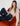 A woman with long brown hair and bangs sits on an orange velvet chair, wearing the Sanctuary Clothing "Fade Into You" ruffle blouse in fawn and high-waisted dark blue jeans, looking at the camera with a relaxed expression.