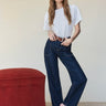 A woman with long light brown hair stands barefoot on a textured carpet, wearing Sanctuary Clothing’s soleil 90s straight jeans in indigo dark raw rinse with a white t-shirt and brown belt, beside a red ottoman against a plain background.