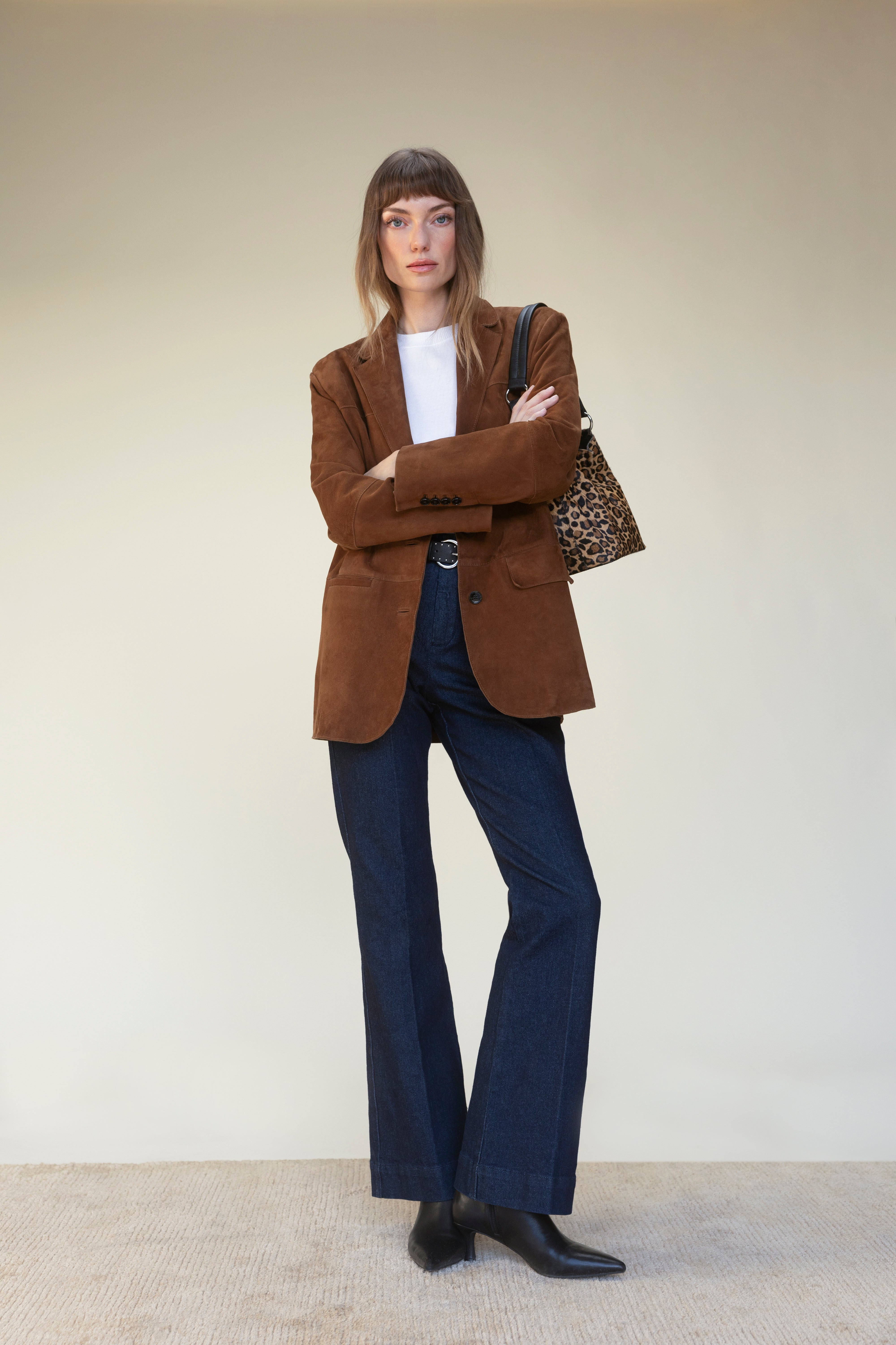 A person stands against a plain background, wearing Sanctuary Clothing's denim stunner jean plot twist with a brown blazer, white shirt, black boots, and a leopard print tote bag, arms crossed and expression neutral.