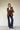 A person stands against a plain background, wearing Sanctuary Clothing's denim stunner jean plot twist with a brown blazer, white shirt, black boots, and a leopard print tote bag, arms crossed and expression neutral.