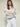 A woman with light brown hair and bangs wears Sanctuary Clothing's boheme popover light oat sweater, paired with white pants and a gray belt, standing confidently with arms crossed against a plain light background.