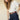 A woman with long brown hair wears the Sanctuary Clothing shirred waist dolman tee in white, tucked into high-waisted dark blue jeans with visible stitching and a brown studded belt, standing against a beige background.