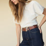 A woman with long brown hair wears the Sanctuary Clothing shirred waist dolman tee in white, tucked into high-waisted dark blue jeans with visible stitching and a brown studded belt, standing against a beige background.