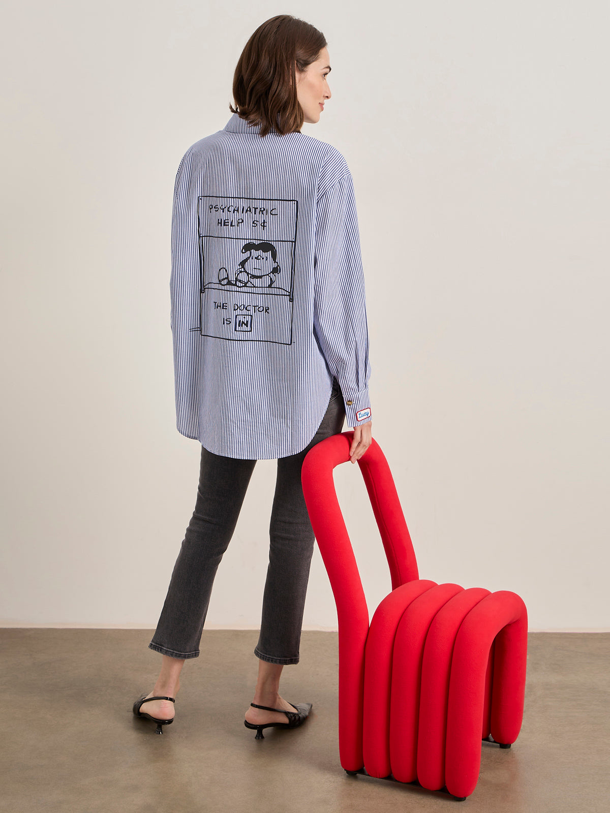 A woman wears the Sanctuary Clothing x PEANUTS 75th Anniversary "the doctor is in shirt blue and white stripe," styled with black jeans and heels, as she stands with a modern red chair in a minimalist room.