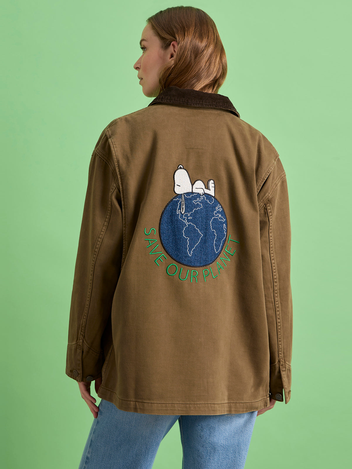 A person wears the "save our planet jacket timber" by Sanctuary Clothing x PEANUTS 75th Anniversary, featuring Snoopy and Woodstock on a globe with “Save Our Planet” text. The background is light green.