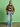 A person stands against a green backdrop, wearing the Sanctuary Clothing x PEANUTS 75th Anniversary "save our planet jacket timber" over a black and white striped shirt, light blue jeans, and beige shoes, with shoulder-length hair and hands in their pockets.