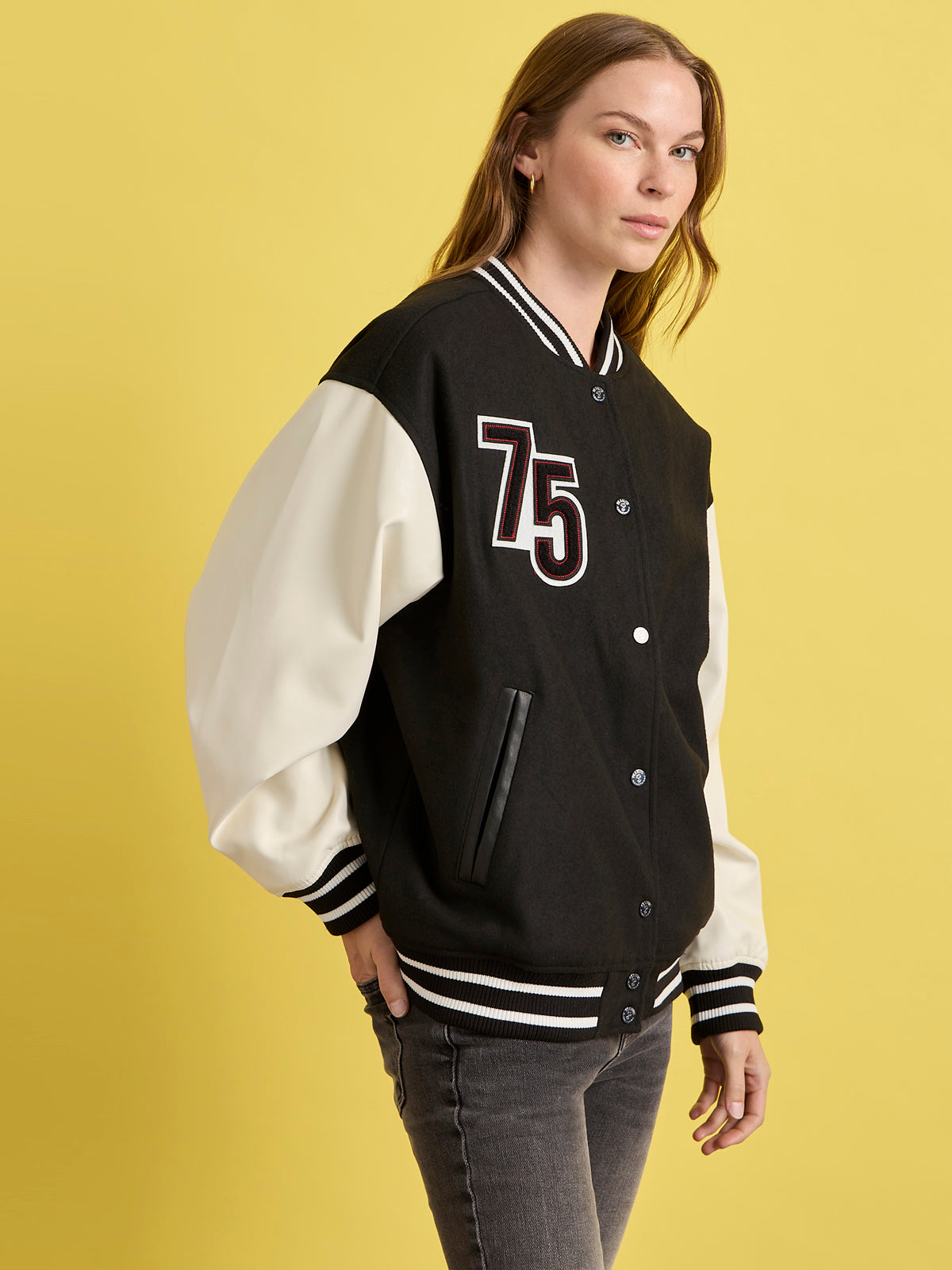 A person models the Sanctuary Clothing x PEANUTS 75th Anniversary "75 varsity jacket black white" against a yellow background, glancing to the side with one hand in their pocket.